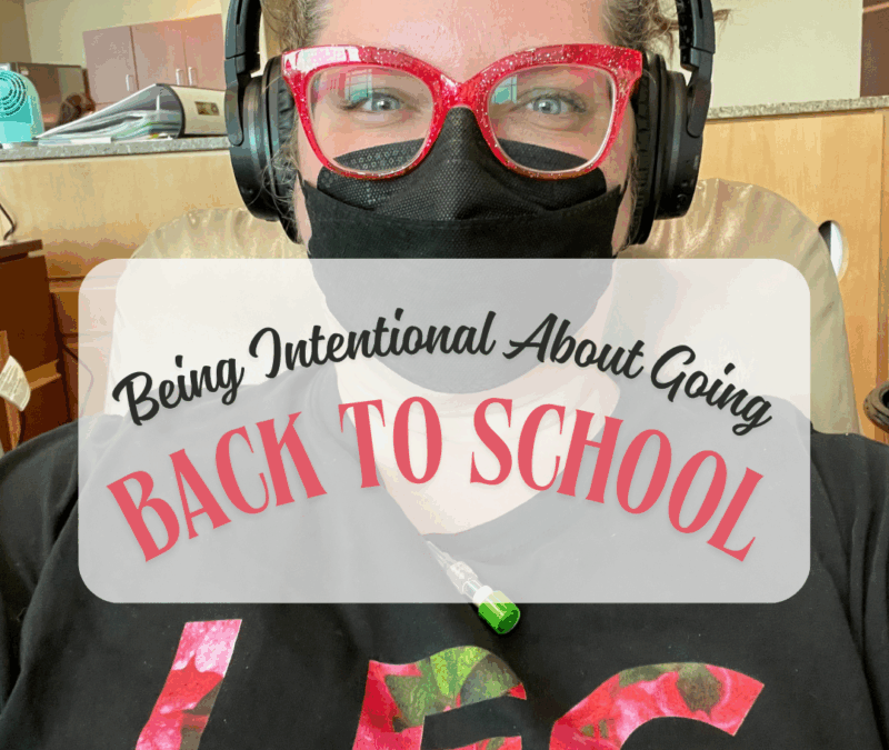 This image shows a person wearing red sparkly glasses, black headphones, and a black face mask, holding up a sign that reads "Being Intentional About Going BACK TO SCHOOL" in decorative lettering. They're wearing a black shirt with colorful letters spelling "LFG" (which commonly stands for "Let's F***ing Go" as an expression of enthusiasm or motivation). The setting appears to be indoors, possibly in an office or institutional building, with modern lighting and furniture visible in the background. The overall tone suggests someone who is excited and motivated about returning to school or an educational environment.